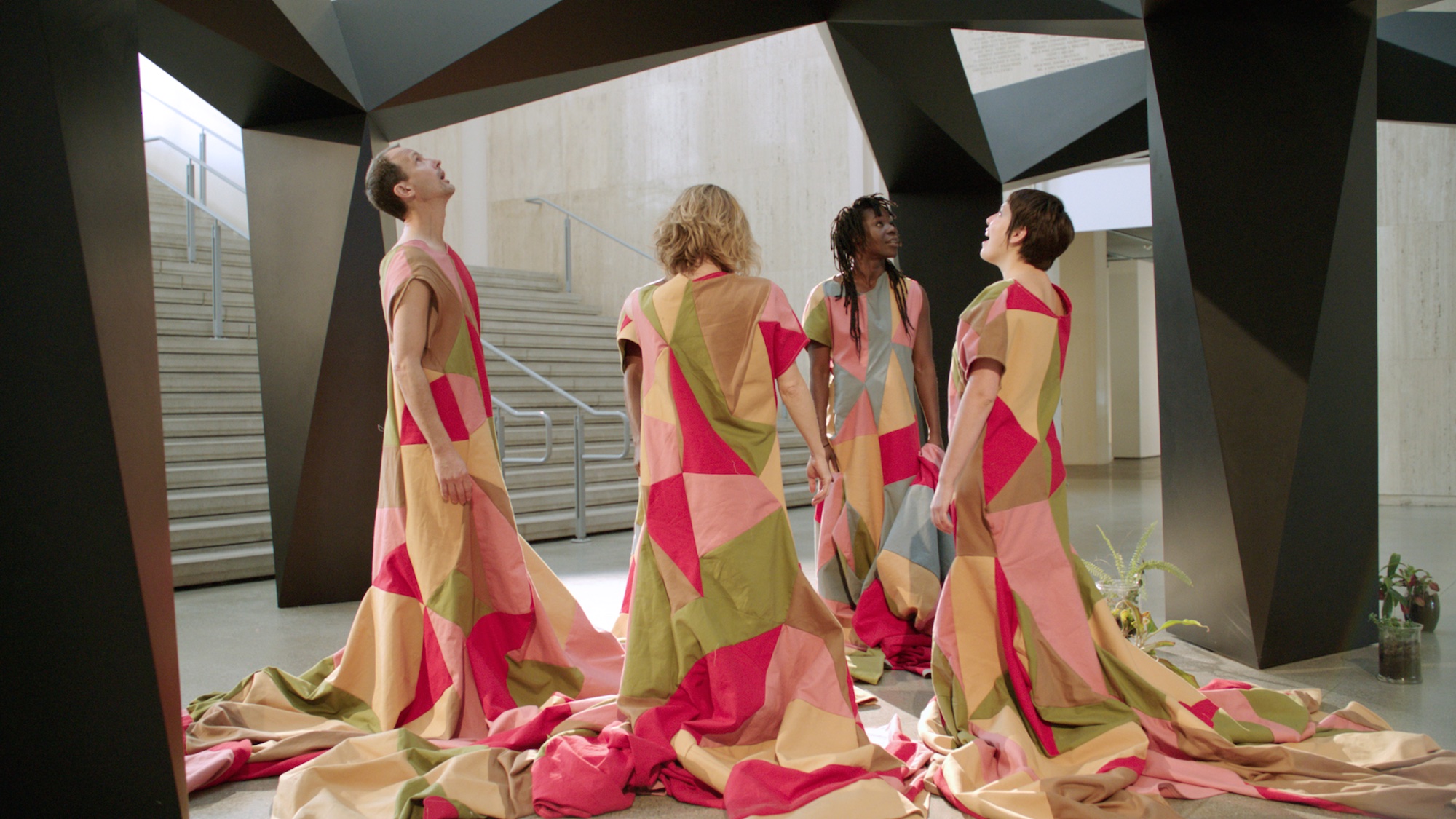 Four performers in long patchwork costumes standing in a circle facing each other within a large black polygonal sculptural structure, appearing to be communicating with it.