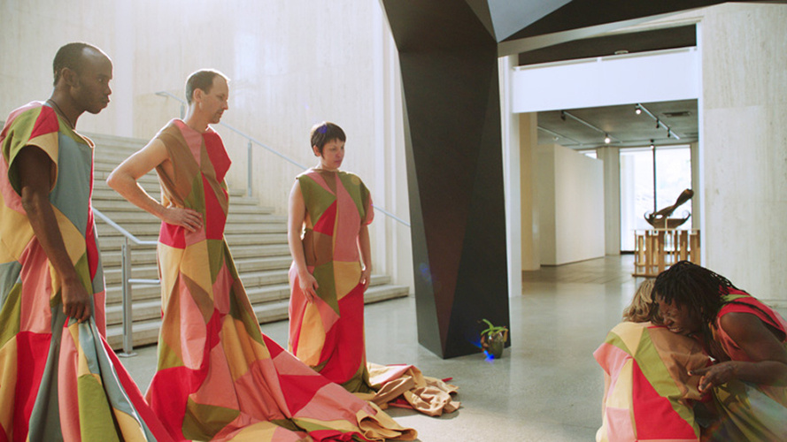 Two performers crouch together on the floor while three others watch; one of the pair appears to be in intense pain or distress.