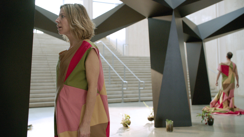 A performer stands closely facing part of a large, black polygonal sculptural structure, appearing entranced by it.