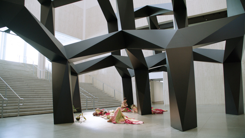 In the distance a group of performers in bright costumes sit and lay on the floor under a large black polygonal sculptural structure inside the vast concrete interior of Los Angeles County Museum of Art