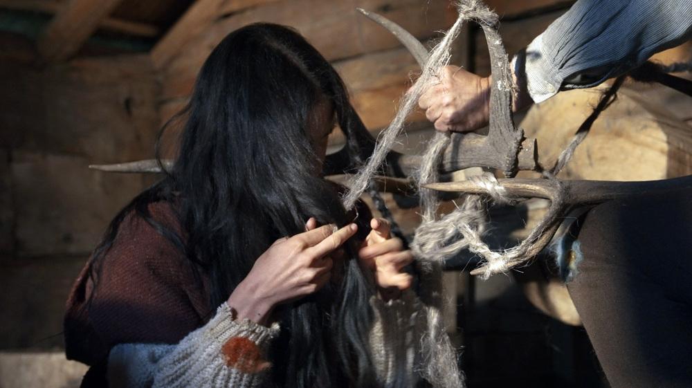 Close-up of a person braiding their dark hair with grey wool or flax tied to antlers or branches, held by an unseen second person
