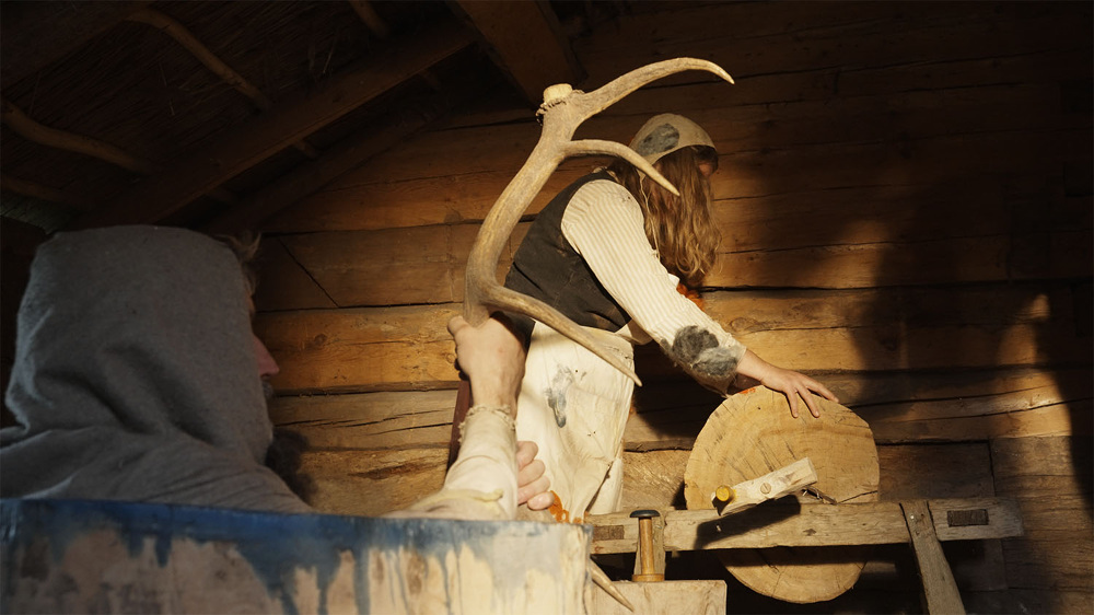 A view inside a rustic wooden cabin featuring a grey-hooded figure from behind holding an antler-shaped object. Further back, a person with long hair and worn clothing operates a large wooden wheel set upon an old trestle.